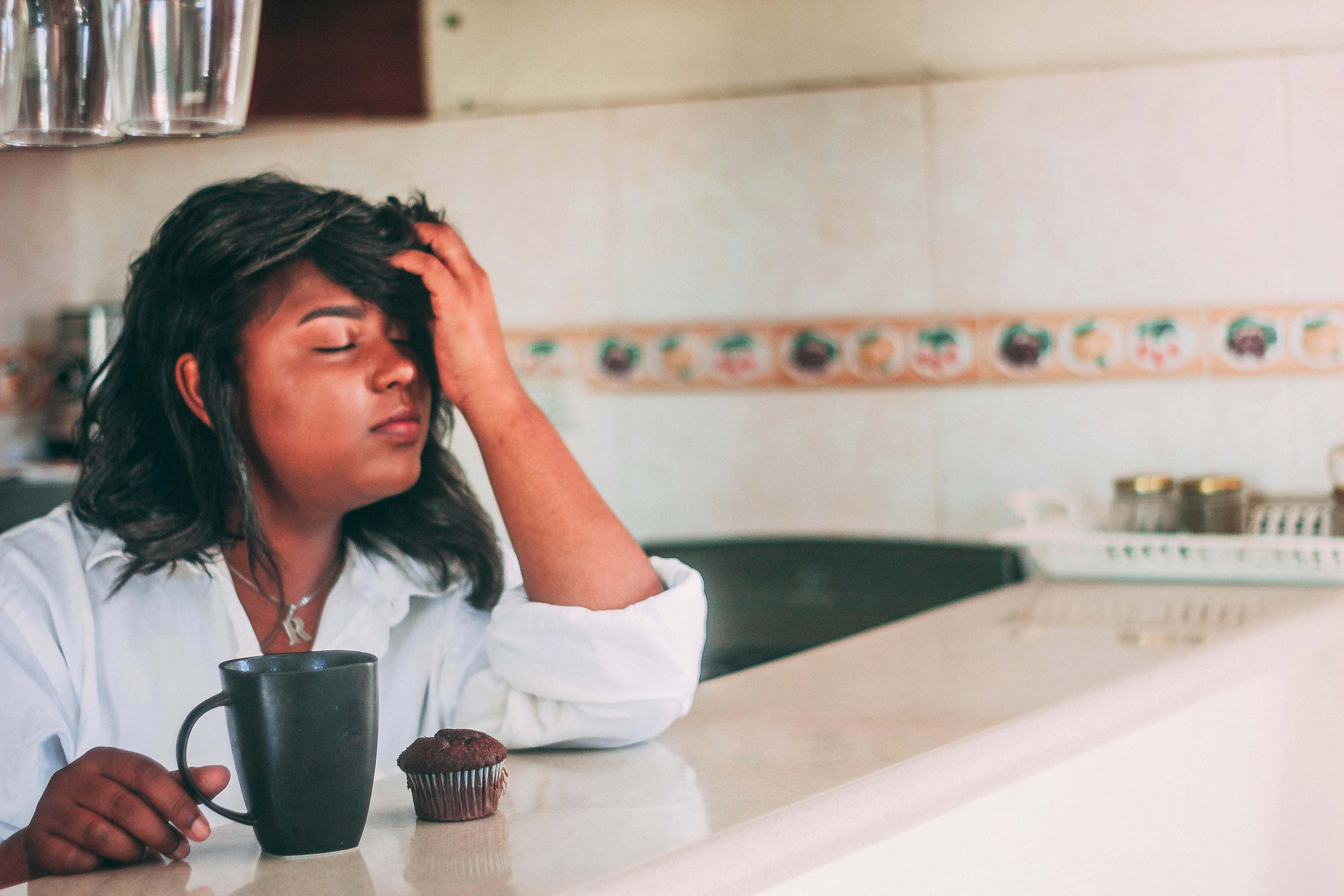 Woman sitting at a kitchen counter with a mug and a muffin, eyes closed, resting her head in her hand, looking exhausted.